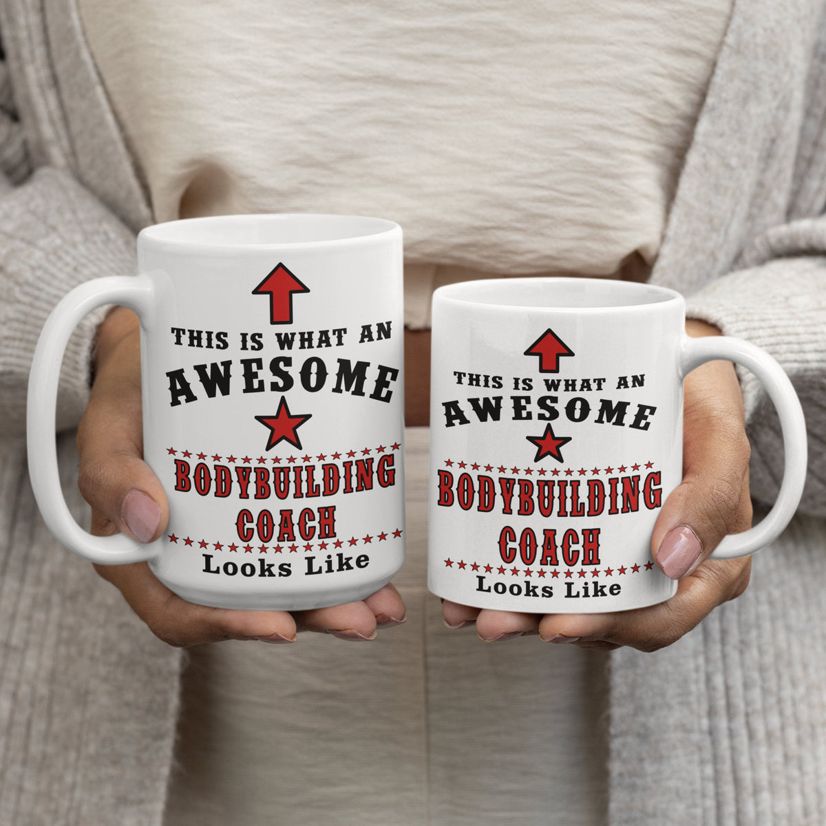 Person holding two white 15oz and 11oz mugs printed with "This Is What An Awesome Bodybuilding Coach Looks Like" in black and red text, comparing mug sizes