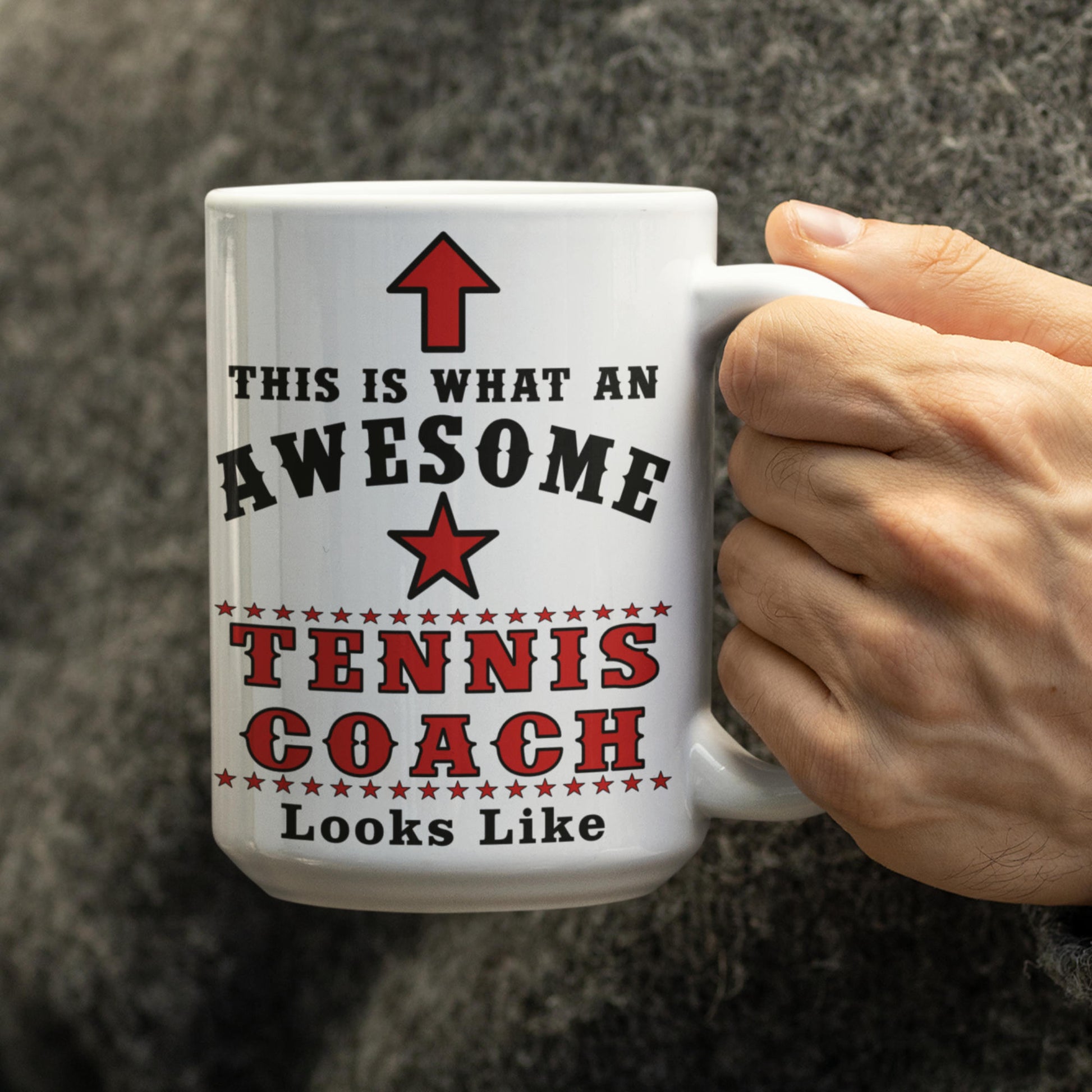 Close-up of a person wearing a grey knitted sweater holding a large 15oz white ceramic mug printed with 'This is what an awesome Tennis Coach Looks Like' in bold red and black.