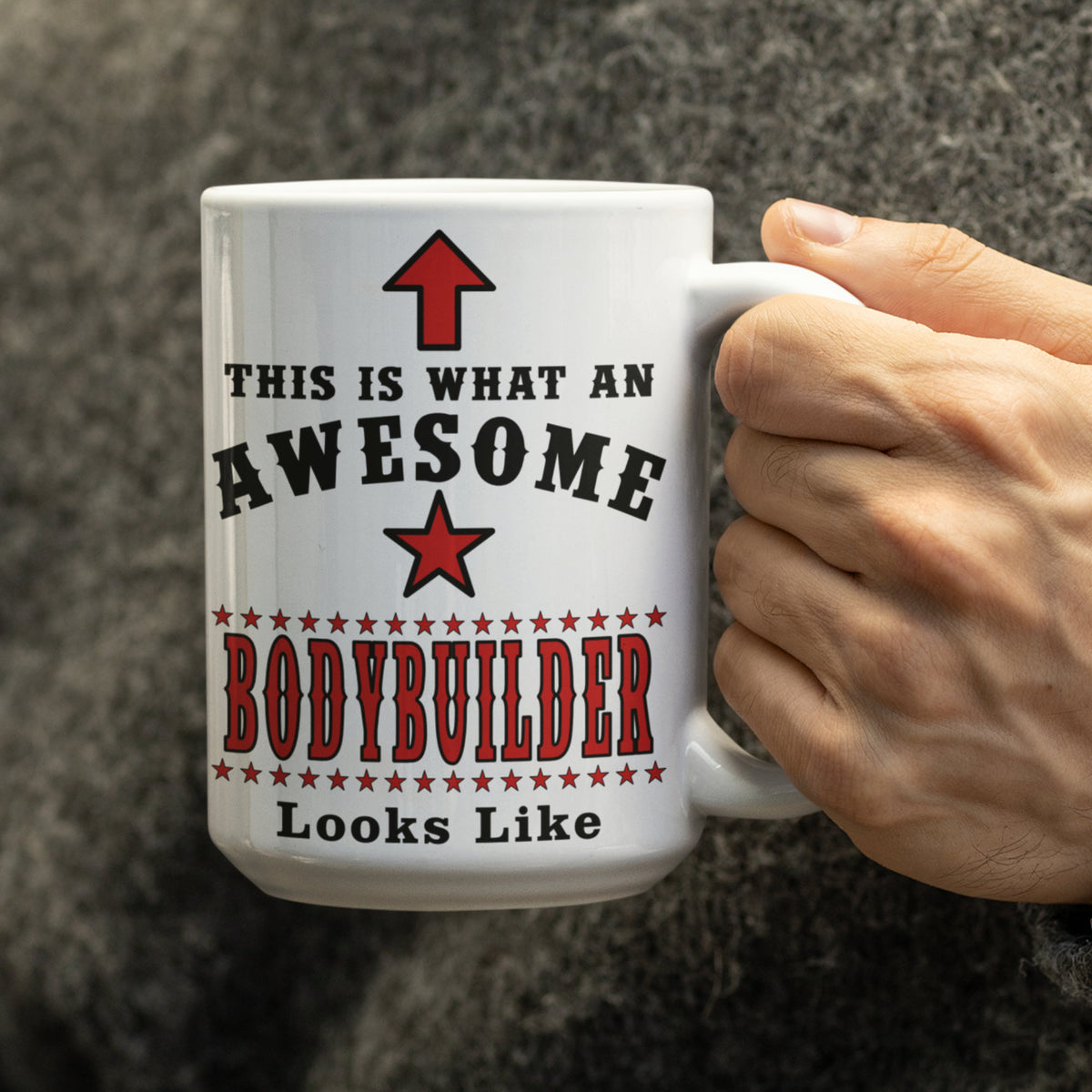 Close-up of a hand holding a white 15oz mug reading "This Is What An Awesome Bodybuilder Looks Like" with red star design
