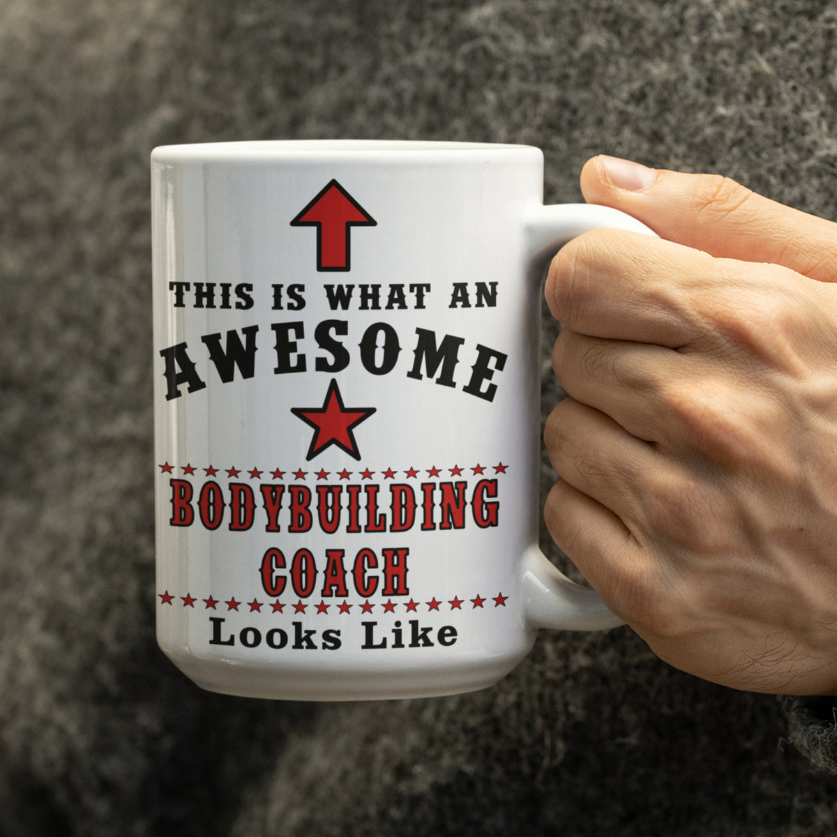 Close-up of hand holding a white 15oz mug with black and red "This Is What An Awesome Bodybuilding Coach Looks Like" text, red star and arrow design