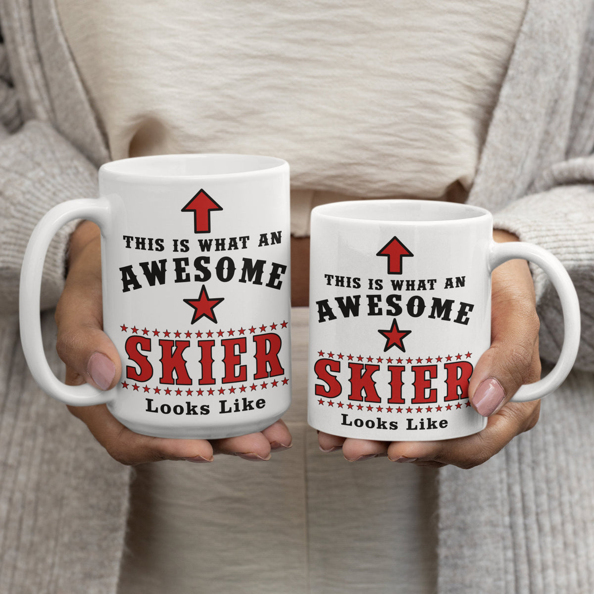 Woman in cream jumper holding two white ceramic mugs printed with "This Is What An Awesome Skier Looks Like" in red and black, comparing 15oz and 11oz sizes